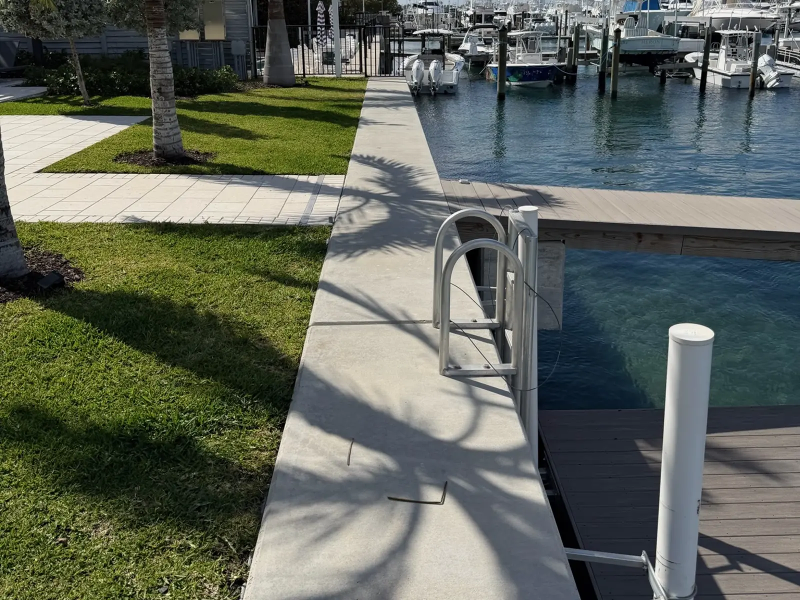 Concrete seawall cap running between a grass lawn and a canal, with a stainless steel ladder, a composite dock, and boats moored at a marina in the background